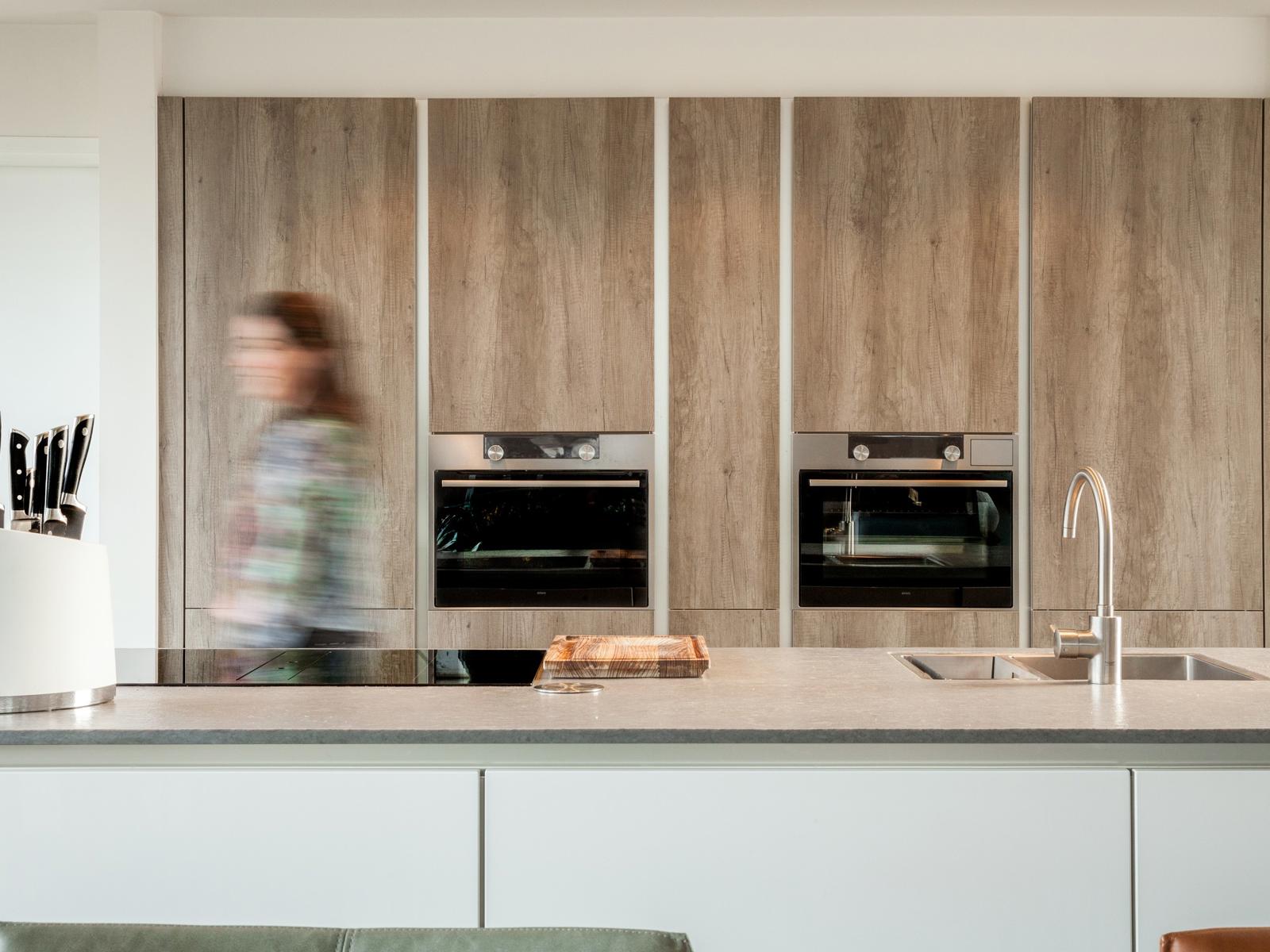 Houtlook keuken in Brunssum: kastenwand in houtlook met twee inbouwovens achter het kookeiland met spoelbak
