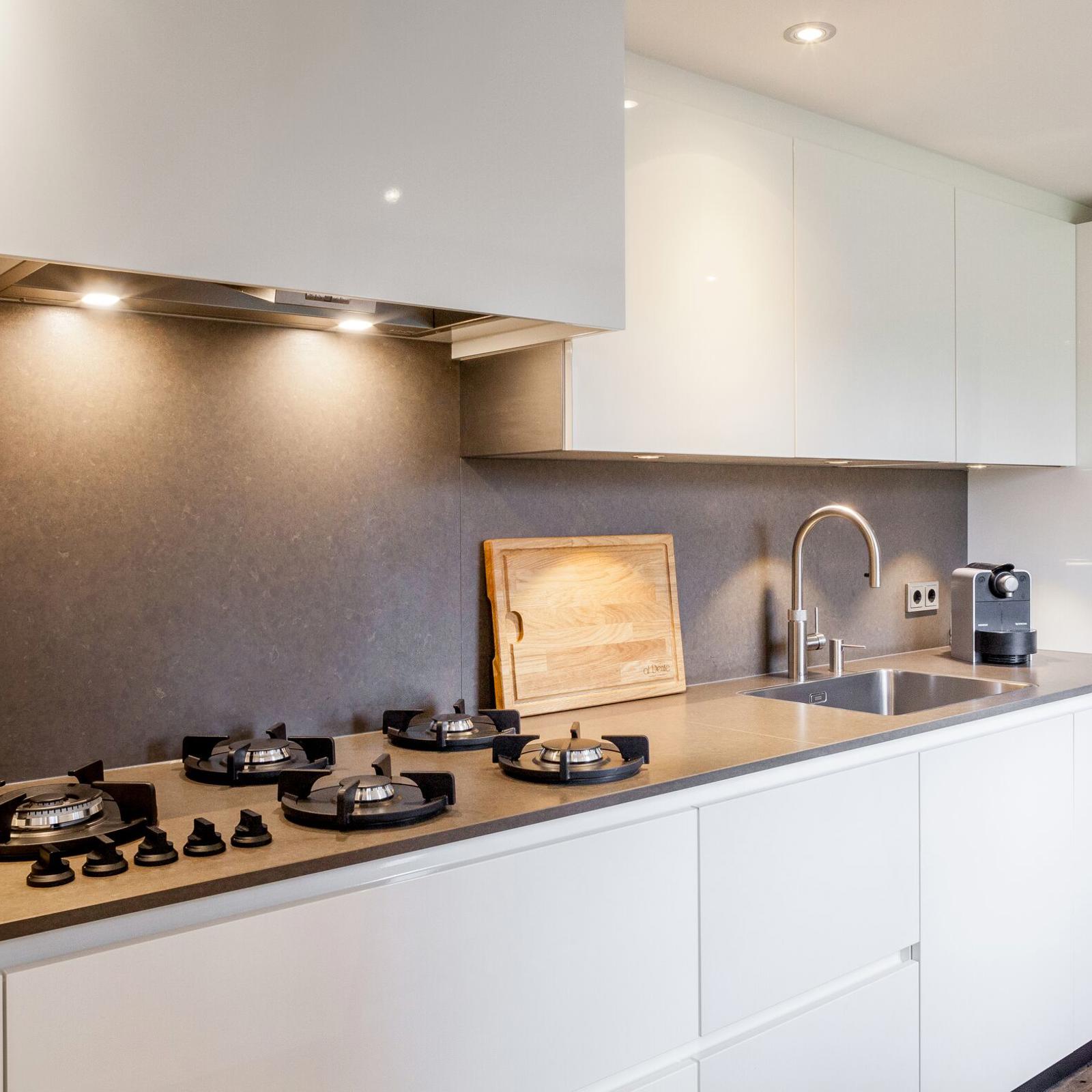 Witte hoogglans keuken met gaskookplaat, taupe achterwand en verlichting onder de bovenkasten