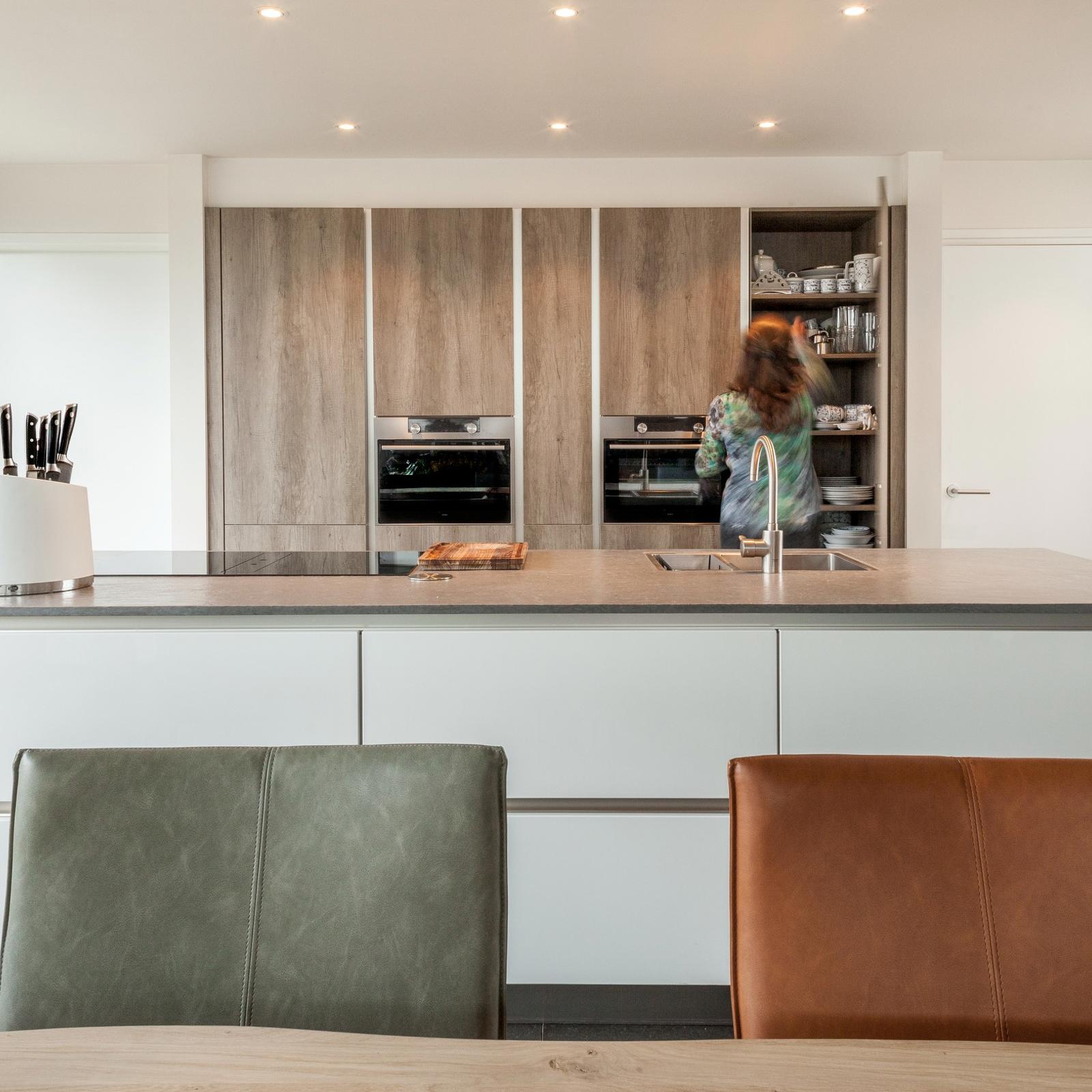 Houtlook keuken in Brunssum: wit kookeiland met eettafel ervoor en een kastenwand in houtlook met open voorraadkast
