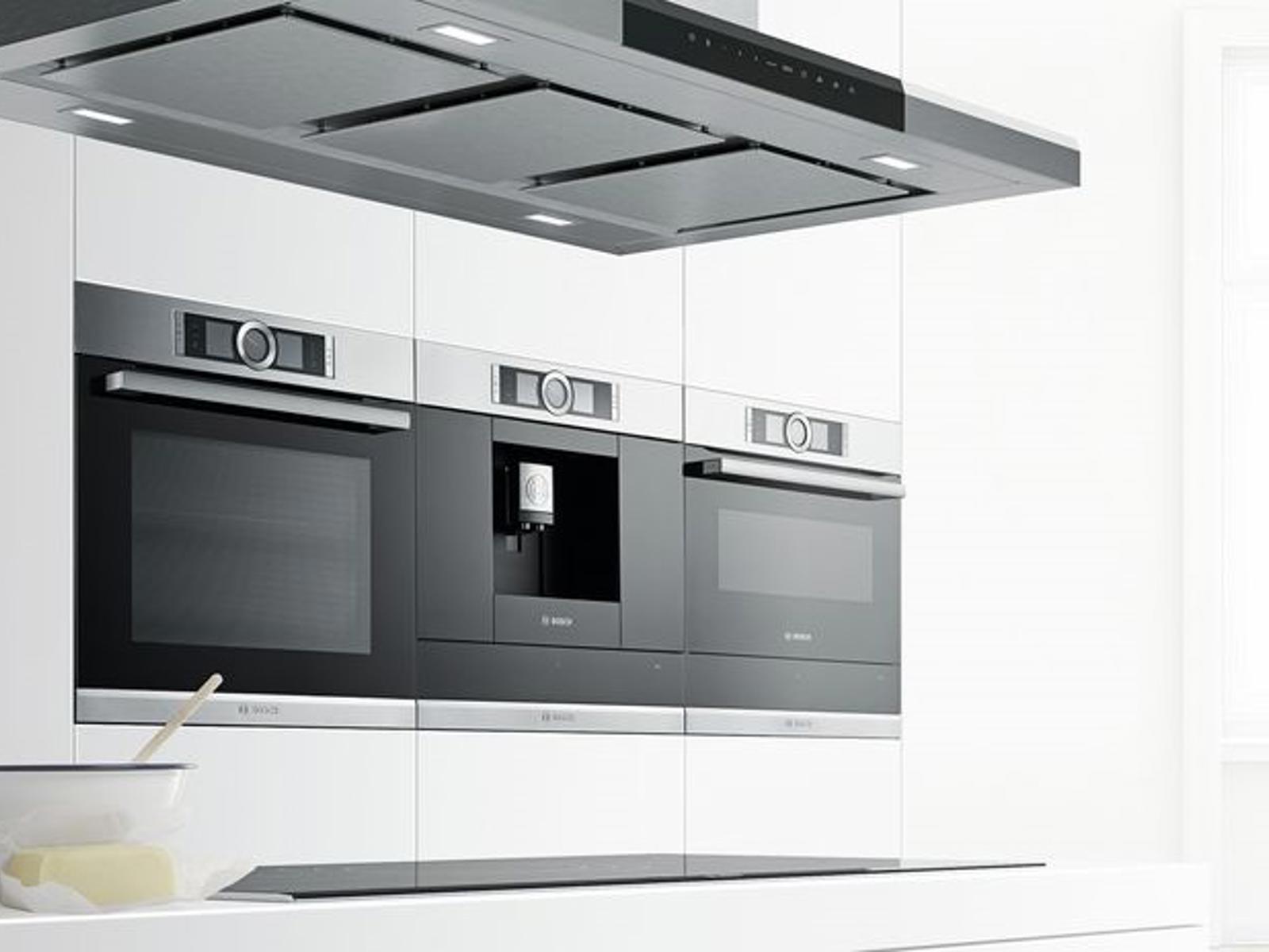Bosch inbouwovens en koffiemachine naast elkaar onder een rvs eilandafzuigkap in een witte keuken